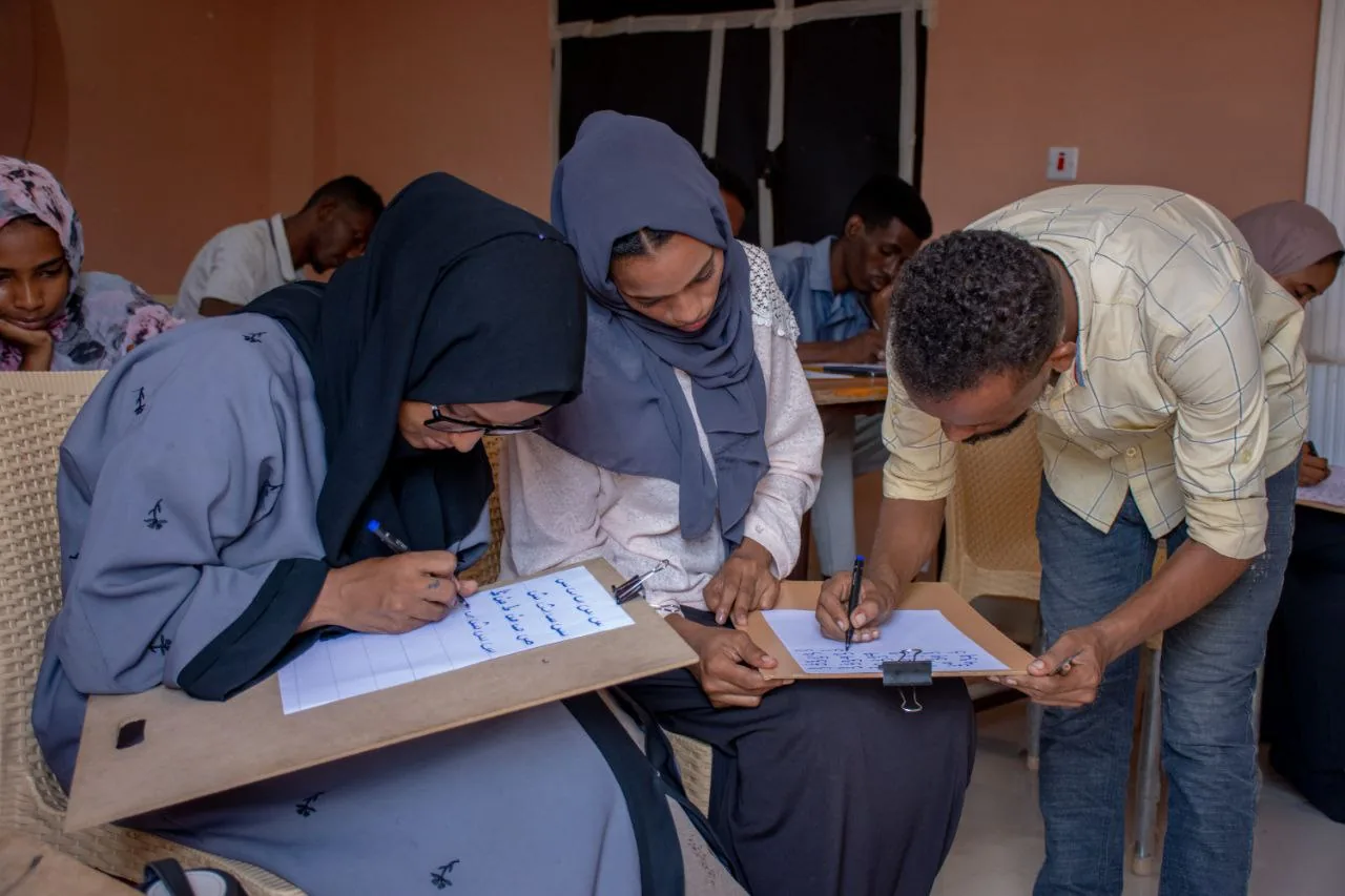 Participants practising Arabic calligraphy at their desks while an instructor leans in to guide them.