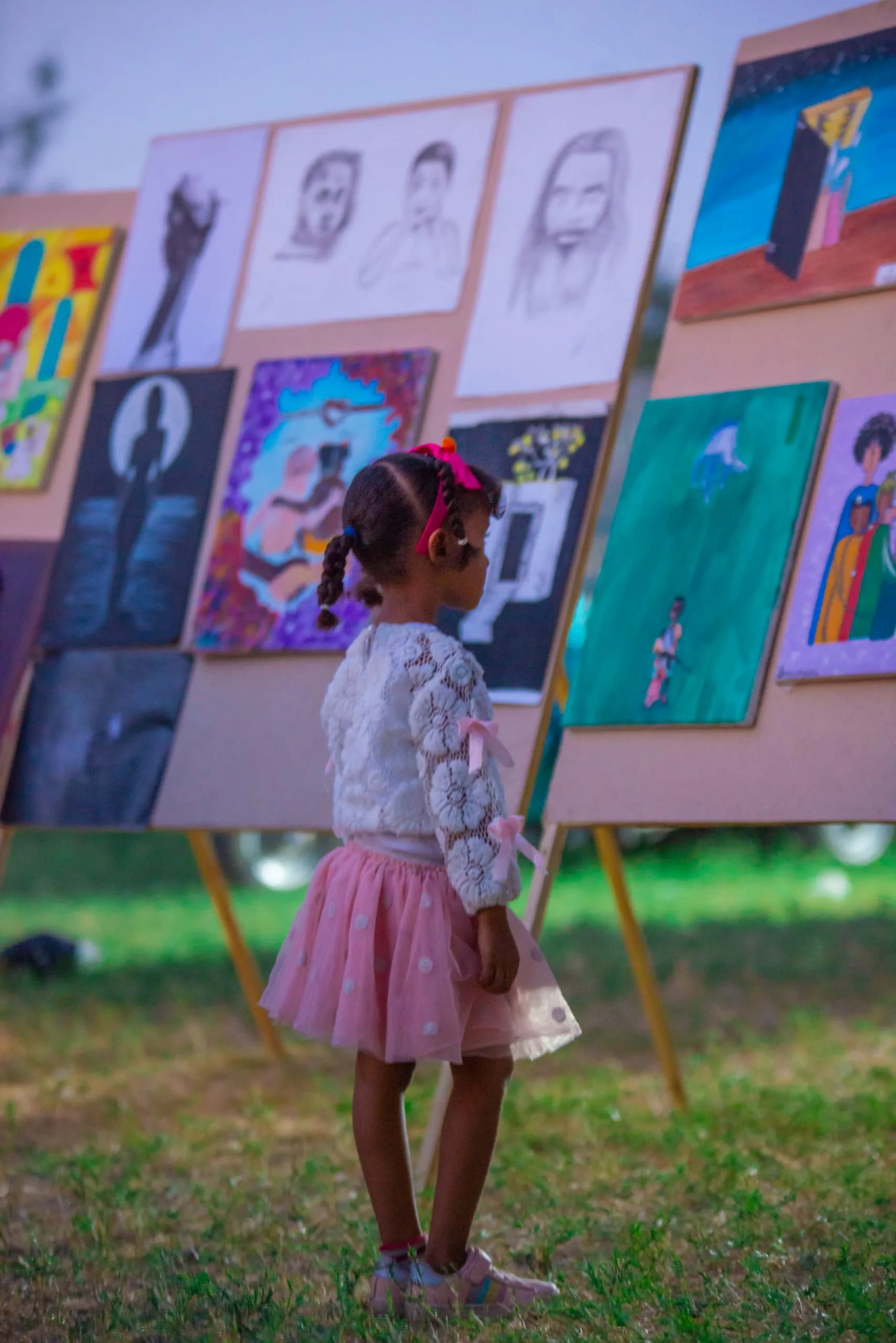 A young girl standing before an open-air wall of paintings at a community exhibition.